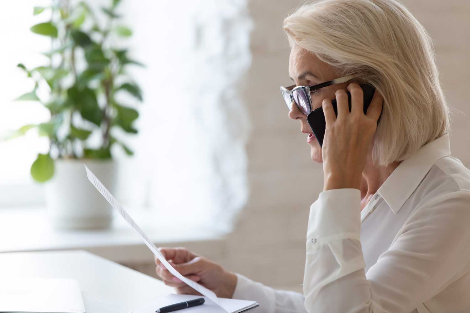 Serious 60 years old businesswoman talking on smartphone and reading document